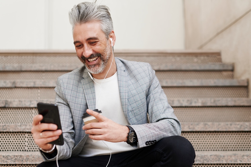 Businessman Sitting Outside In Video Call Laughing mature businessman drinking a takeaway coffee and doing a video call on his smartphone while sitting on steps in the city