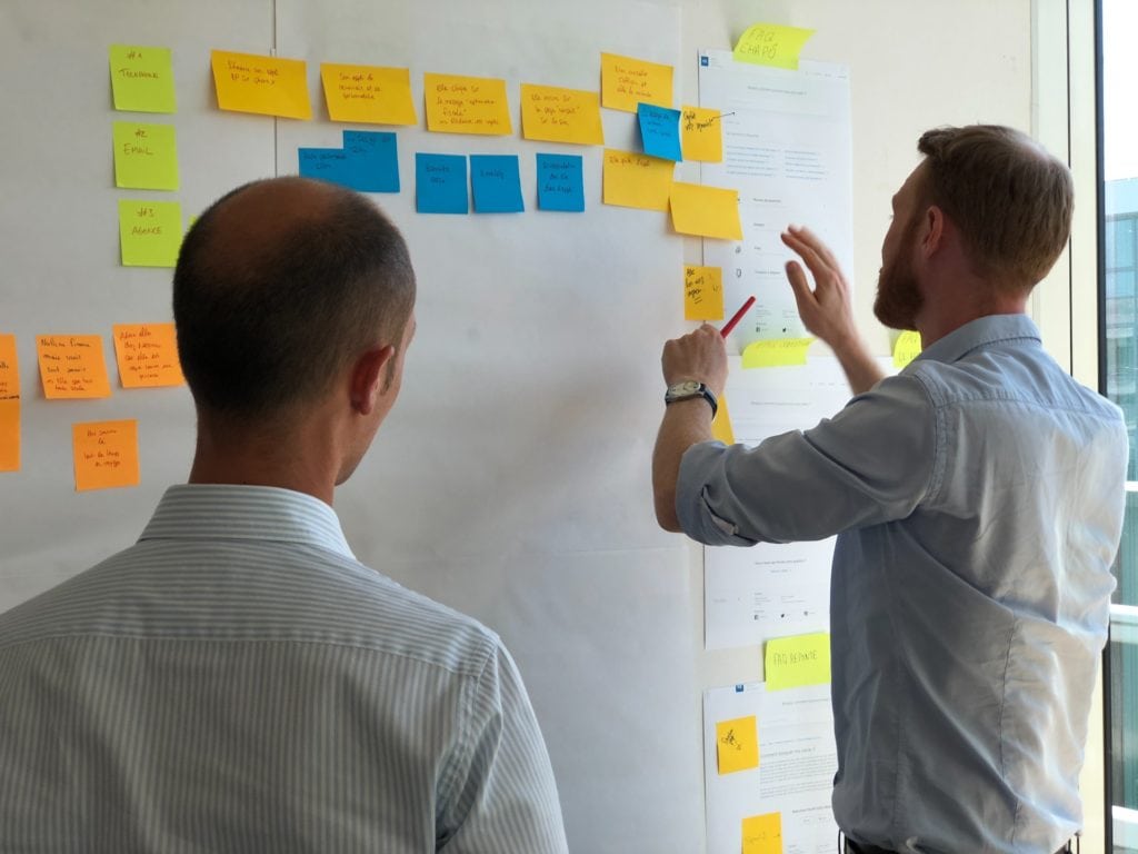 Two Men at Workplace Pinning Post-It Notes on WhiteBoard 