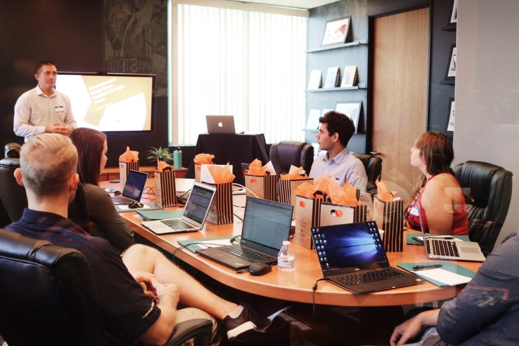 Man Standing In Front of Screen Addressing Team Sitting Across Boardroom Table